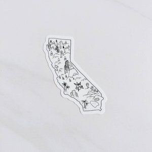May include: A black and white sticker in the shape of California. The sticker features a detailed outline of the state with illustrations of mountains, trees, a desert landscape, and buildings. The sticker is on a white background.