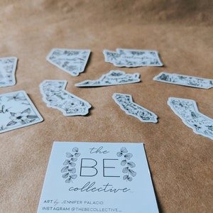 May include: A collection of black and white state-shaped stickers with floral designs, scattered on a brown surface. A business card with the text "the BE collective" and contact information is in the foreground.
