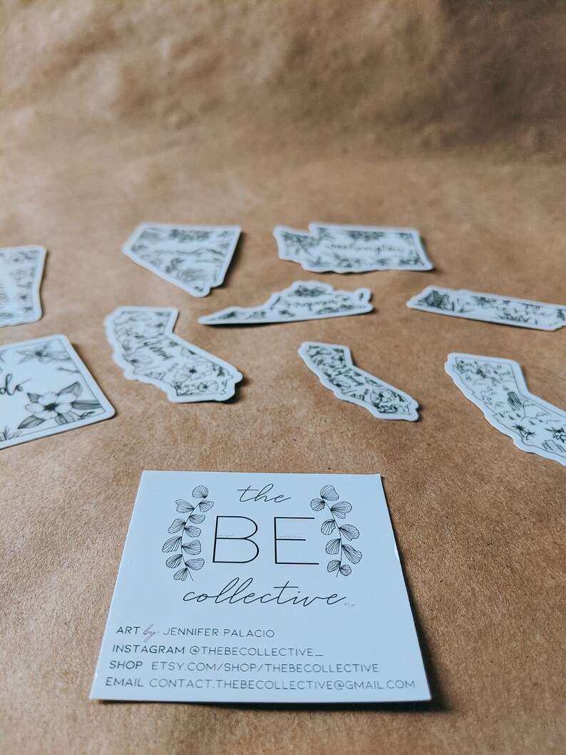 May include: A collection of black and white state-shaped stickers with floral designs, scattered on a brown surface. A business card with the text "the BE collective" and contact information is in the foreground.