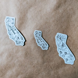 May include: Three white stickers in the shape of California. Each sticker features a black outline of the state with different designs: floral, botanical, and landscape illustrations. The word "California" is written on two of the stickers.