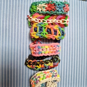 May include: A stack of colorful rainbow loom bracelets, including a black and rainbow bracelet, a pink and white bracelet, a white and rainbow bracelet, a blue and green bracelet, and a green and white bracelet.