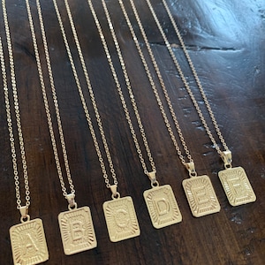 May include: Seven gold necklaces with square pendants featuring the letters A through G. The pendants have a textured, raised design.