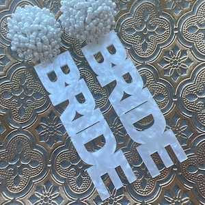 May include: White beaded earrings and matching "BRIDE" word cutouts. The earrings are round and fluffy, while the word cutouts have a pearlescent finish. The items are displayed on a gold and silver patterned surface.