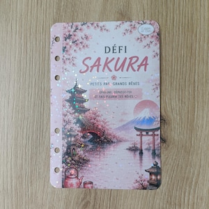 Può includere: Un'agenda rosa e bianca con il testo "DÉFI SAKURA" e illustrazioni in stile giapponese. L'agenda presenta una pagoda, un ponte e il Monte Fuji, con fiori di ciliegio. Il testo "PETITS PAS, GRANDS RÊVES" è visibile.