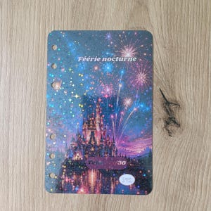 May include: A notebook cover with a fantasy castle scene, fireworks, and the text "Féerie nocturne." The cover has a blue and purple color scheme with a glittery effect. It has several holes for binding.