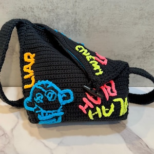 May include: Black crocheted bag with a colorful cartoon monkey, and the words "Liar", "High", "Black", and "Even" in neon pink, yellow, and blue yarn.