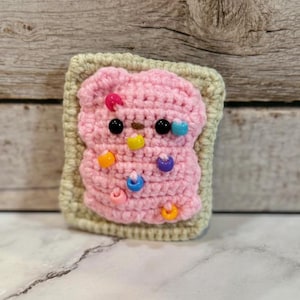 May include: A handmade crochet pop-tart in a square shape. The pop-tart has a pink frosting with colorful bead sprinkles and black bead eyes. The pop-tart is surrounded by a light green border.