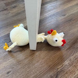 May include: A crocheted chicken toy with yellow beak, red wattle, and white body, stuck under a white door frame.