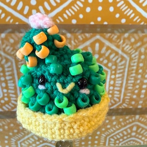 May include: A green crocheted cactus with a pink flower on top. The cactus is decorated with green and orange beads.