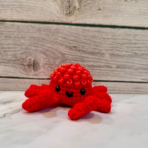 May include: A red crocheted crab with black eyes and a smiling mouth. The crab has a textured shell and is sitting on a white surface.