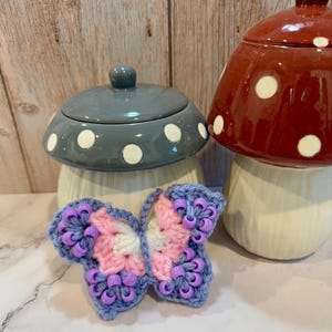 May include: A crocheted butterfly with purple and pink yarn and purple beads. The butterfly is sitting on a white surface in front of two ceramic mushroom containers with white polka dots.