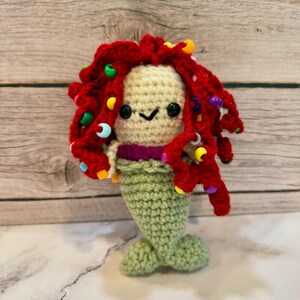 May include: A crocheted mermaid doll with red yarn hair and colourful beads. The doll has a green tail and a smiling face.