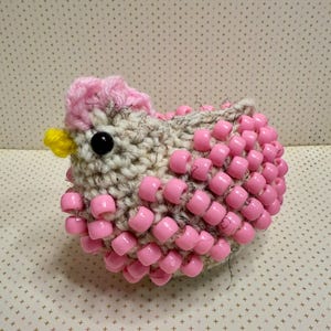 Peut inclure: Un jouet en crochet en forme de poussin avec des perles roses et un pompon rose sur la tête.
