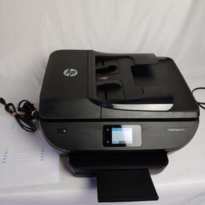May include: Black HP ENVY Photo 7858 all-in-one printer. The printer has a touchscreen display and a paper output tray. The HP logo is visible on the top left. A power cord and a sheet of paper are next to the printer.