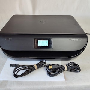 May include: Black HP ENVY 4520 all-in-one printer with a small LCD screen and power cord. The printer has a sleek, modern design. The power cord and USB cable are neatly bundled. The printer is on a white surface.