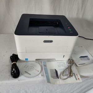 Puede incluir: Una impresora Xerox blanca y azul marino con un cable de alimentación, un CD y manuales. La impresora tiene una bandeja de papel y un panel de control. Los accesorios incluyen un cable USB y un cable de alimentación.