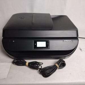 May include: Black HP ENVY 5030 all-in-one printer with a small display screen. The printer has a flat top with a hinged lid and a curved front. Two black power cords are bundled with rubber bands.
