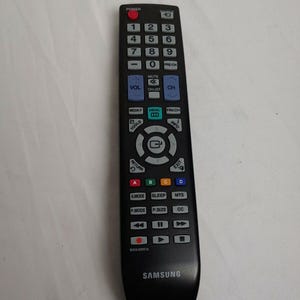 Könnte beinhalten: Schwarze Samsung TV-Fernbedienung mit zahlreichen Tasten, darunter Ein/Aus, Lautstärke, Kanal und Zifferntasten. Die Fernbedienung hat ein geschwungenes Design und das Samsung-Logo unten.