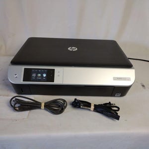 May include: A black and silver HP ENVY 5660 all-in-one printer with a touchscreen display. The printer has a flatbed scanner and is accompanied by two black power cords.