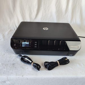 May include: A black HP printer with a small display screen and the HP logo on top. Two black power cords are bundled with rubber bands. The printer sits on a white surface.