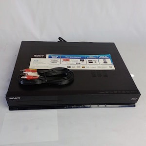 Könnte beinhalten: Schwarzer Sony Blu-ray-Player mit einem Satz roter, weißer und gelber Cinch-Kabel. Der Player hat einen Aufkleber mit Funktionen und Spezifikationen. Der Player befindet sich auf einer weißen Oberfläche.