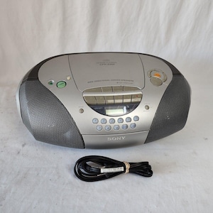 Può includere: Boombox Sony argento e nero con lettore CD, lettore di cassette e radio AM/FM. Il boombox ha un display digitale, più pulsanti e due altoparlanti. Un cavo di alimentazione nero è di fronte al boombox.