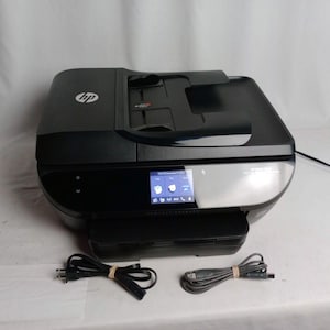 May include: A black and gray HP ENVY Photo 7855 all-in-one printer with a touchscreen display. The printer has a paper tray and a scanner. Included are a power cord and a USB cable.