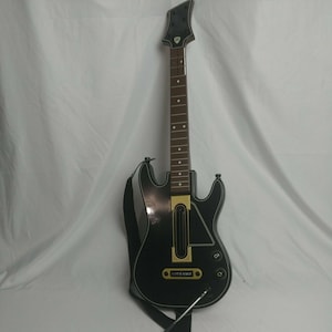 Pode incluir: Um controlador de jogo preto em forma de guitarra com uma alça preta. O controlador tem um braço marrom com incrustações de pontos brancos e um cabeçote preto. O texto "LET'S POWER" é visível no corpo.
