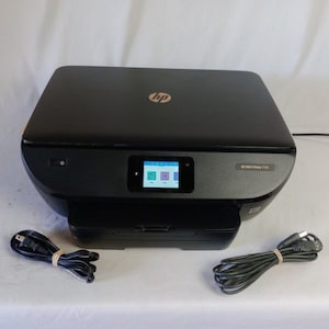May include: A black HP ENVY Photo 7155 all-in-one printer with a small color screen and a paper tray. The printer has a power cord and a USB cable. The HP logo is visible on the top.
