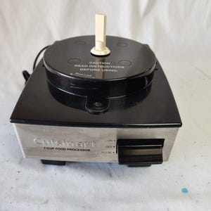Puede incluir: Una base de procesador de alimentos Cuisinart de 7 tazas con una parte superior negra y un panel frontal de acero inoxidable. La parte superior tiene un eje central blanco y el texto CAUTION READ INSTRUCTIONS BEFORE USING. Incluye un interruptor negro.