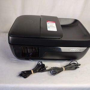 May include: Black HP OfficeJet 3830 all-in-one printer. The printer has a flatbed scanner and a paper tray. Two black power cords are bundled with rubber bands. The printer is on a white surface.