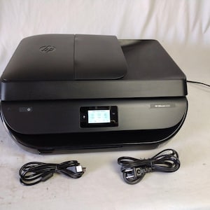 May include: Black HP OfficeJet 5255 all-in-one printer with a small LCD screen and two black cords. The printer has a flatbed scanner and a top cover with the HP logo. The printer is on a white surface.