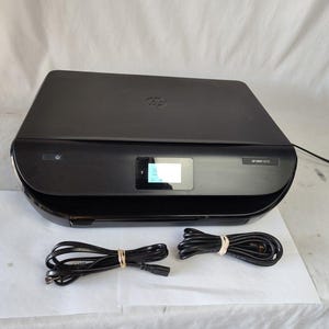 May include: A black HP ENVY 5012 all-in-one printer with a small display screen. Two black power cords are bundled with rubber bands. The printer is on a white surface.