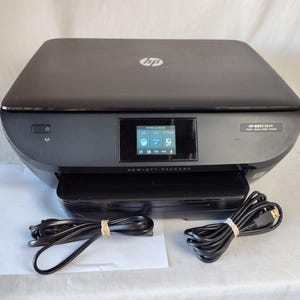 Puede incluir: Una impresora multifunción HP ENVY 5645 negra con una pequeña pantalla LCD que muestra iconos. La impresora tiene una bandeja de papel y viene con un cable de alimentación y un cable USB. El logotipo de HP es visible en la parte superior.