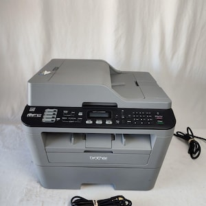 May include: A gray Brother MFC-L2710DW multifunction laser printer featuring a top document feeder, a central control panel with a digital display, a numeric keypad, and function buttons. The unit includes a paper tray and a black power cord.