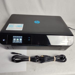 May include: Black HP all-in-one printer with a small LCD screen and a blue HP logo. The printer has a glossy finish and includes two bundled black cables. A sticker on the top reads "50% + FREE".