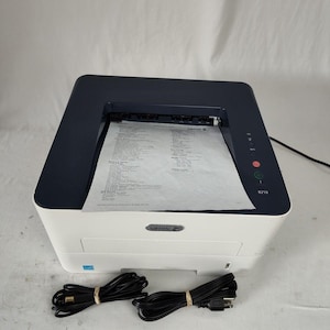 Puede incluir: Una impresora Xerox 8210 blanca y azul marino con un documento impreso en la bandeja de salida. Dos cables de alimentación negros están delante de la impresora. La impresora tiene una pegatina azul Energy Star.