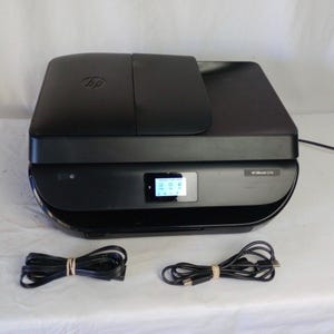 May include: A black HP ENVY 5275 all-in-one printer with a small LCD screen and two bundled power cords. The printer has a sleek, modern design and is set against a white background. The HP logo is visible on the top.