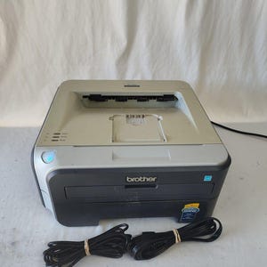 May include: A Brother brand laser printer with a black and gray design. The printer has a paper output tray and a power cord. The printer is on a white surface.