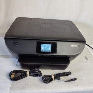 May include: A black HP ENVY 5660 wireless printer with a small LCD screen displaying icons. Two black USB cables are included. The printer is on a white surface.