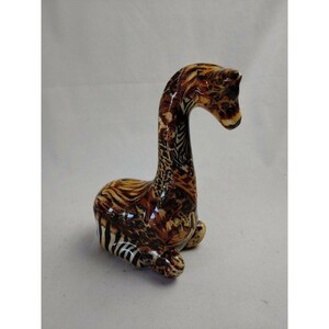 May include: A ceramic giraffe figurine in a seated position. The giraffe has a long, curved neck and is decorated with a brown and tan animal print pattern. The figurine is a decorative item.