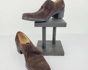 May include: A pair of brown suede heeled shoes with a buckle strap. The shoes are on a wooden display stand.