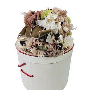 Puede incluir: Una caja redonda blanca con un borde rojo y una cuerda roja atada alrededor. La caja está llena de una variedad de flores secas en tonos de marrón, blanco y verde.