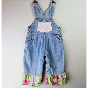 May include: Blue denim overalls with floral print trim. The overalls have a pink floral patch pocket on the front.