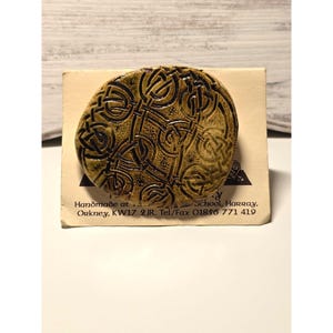 Puede incluir: Broche ovalada de cerámica hecha a mano con un diseño de nudo celta en tonos marrón y beige. La broche se muestra sobre una tarjeta crema con el texto: "Handmade at... Orkney, KW17 2JR. Tel/Fax 01856 771 419."