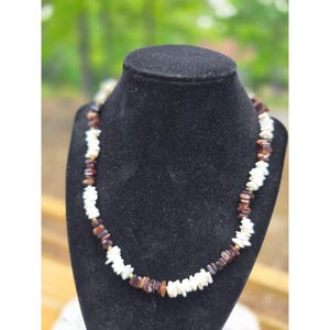 Puede incluir: Un collar expuesto sobre un busto negro. El collar presenta una mezcla de cuentas blancas, marrones y doradas. Las cuentas están dispuestas en un patrón, con las blancas intercaladas con las marrones y doradas. Es una pieza de joyería.