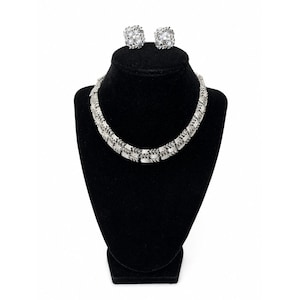Puede incluir: Un collar plateado y un conjunto de pendientes cuadrados a juego. Las joyas se exhiben sobre un busto de terciopelo negro. El collar y los pendientes presentan piedras transparentes de corte cuadrado.
