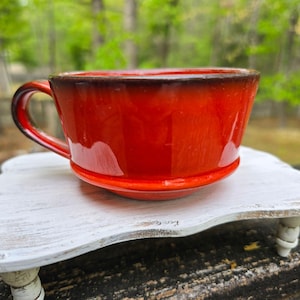 Puede incluir: Una taza de té de cerámica roja brillante con una base de platillo a juego y un asa curva. El borde de la taza tiene un esmalte marrón oscuro. La taza está sobre una bandeja de madera blanca envejecida, con un fondo verde borroso.