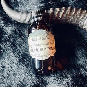May include: A dark brown glass bottle with a label that reads "Sister of Salem" and "Moon Water". The bottle is sitting on a black fur surface.