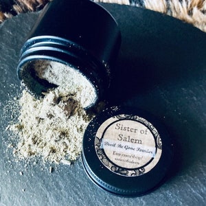 May include: A black jar with a label that reads "Sister of Salem, Devil Be Gone Powder, Etsy.com/shop/sisterofsalem". The jar is open and the contents are spilling out. The contents are a light brown powder.
